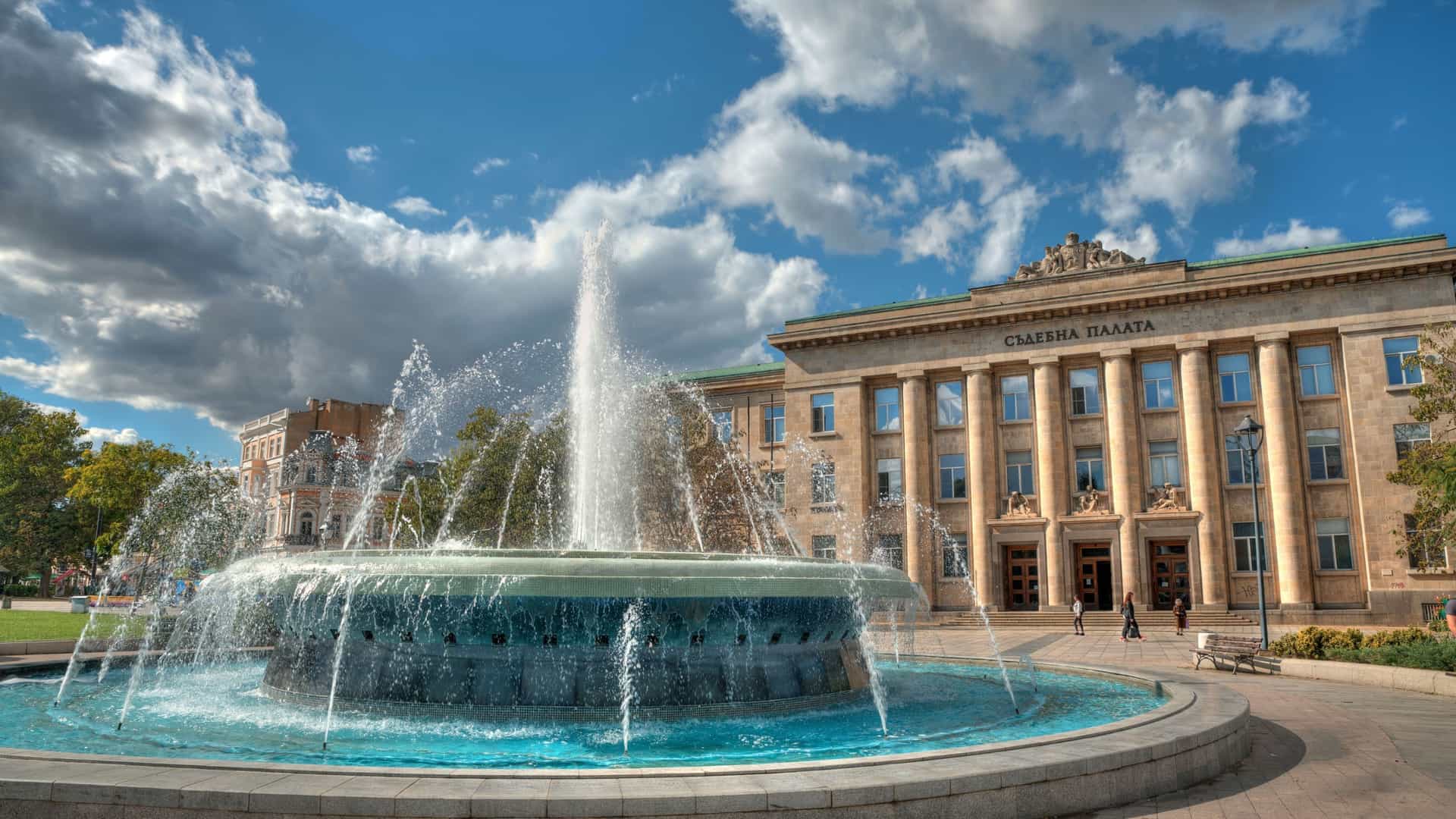 A vibrant photograph of Freedom Square in Ruse, Bulgaria, featuring a large, ornate fountain spraying water in the foreground, with the majestic Courthouse building and a bright blue sky behind it.