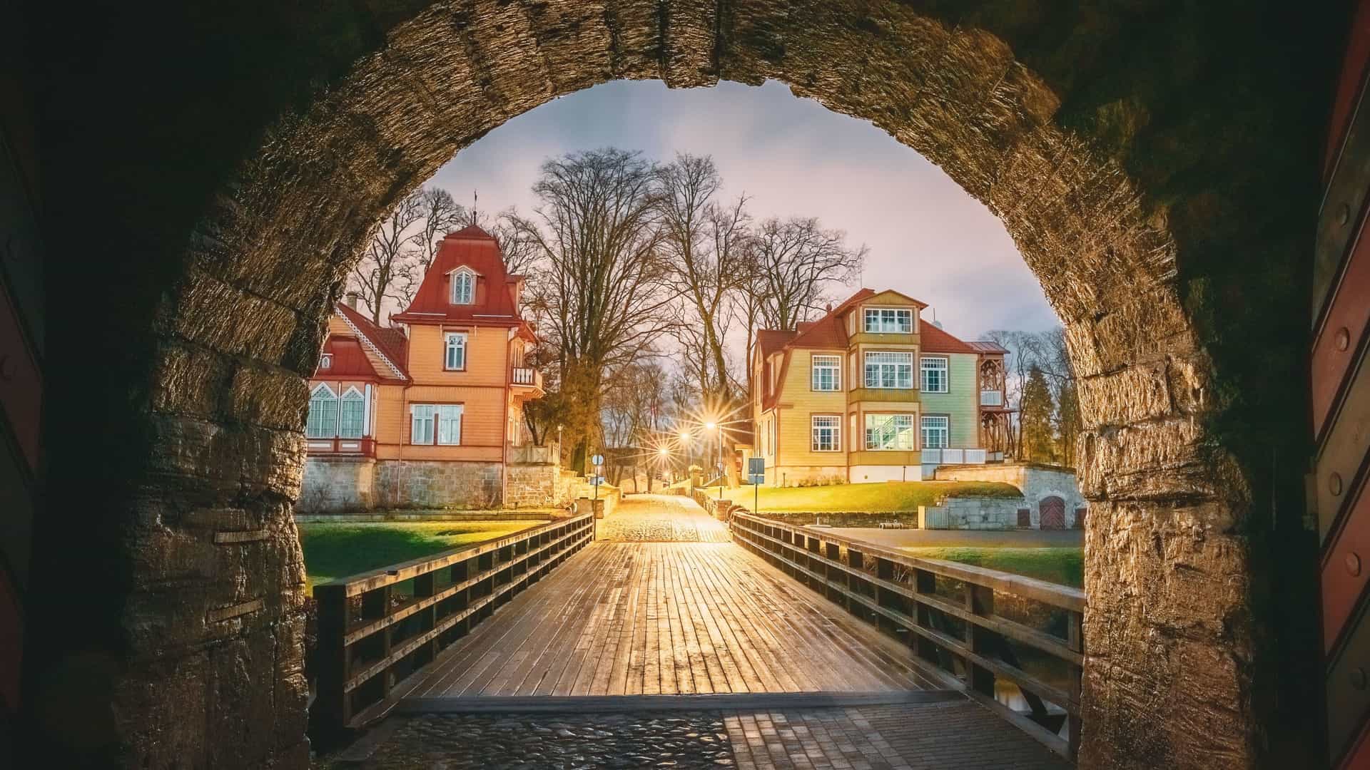 A beautiful view through a stone archway leading to a wooden bridge over a moat, with historic, brightly colored houses on a cobblestone street at dusk in Kuressaare, Saaremaa, Estonia.