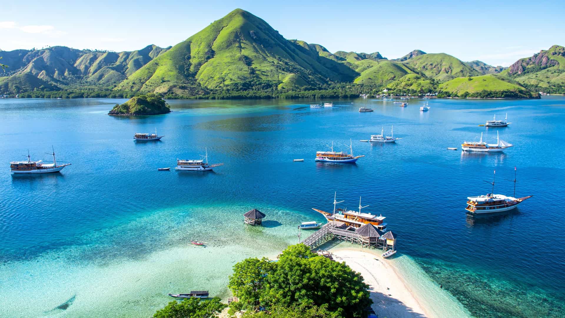 Traditional wooden boats anchor in the turquoise bay near a small beach, framed by steep, grassy green hills on Sabalana Island, Indonesia.