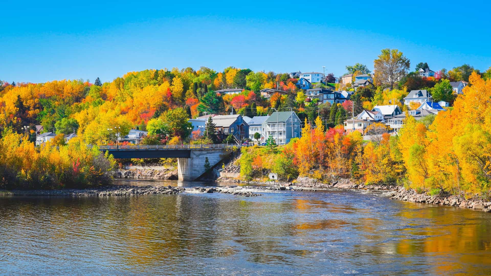 A beautiful autumn landscape in Saguenay with a river flowing under a bridge, surrounded by hillsides adorned with houses and vibrant yellow, orange, and red fall foliage.