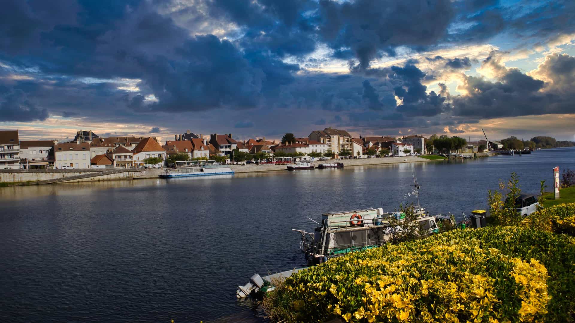 A beautiful sunset view of the Saône River in Saint-Jean-de-Losne, France, with a row of traditional houses and boats docked along the bank under a dramatic, cloudy sky.