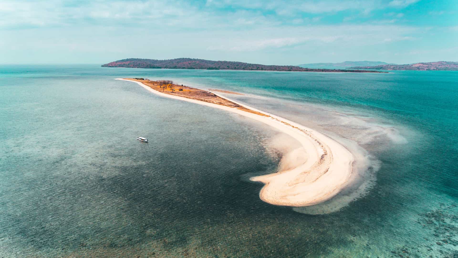 A high-angle aerial view of a long, narrow white sandbar curved like a crescent in the turquoise waters of Saleh Bay, Sumbawa, Indonesia, with a lone boat anchored nearby.