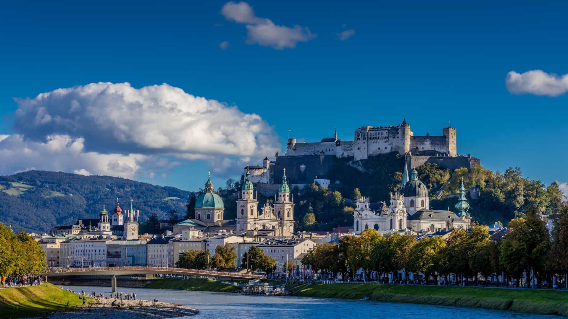 A picturesque view of the historic city of Salzburg, Austria, with the Salzach River in the foreground, the Hohensalzburg Fortress perched on a hill, and a beautiful mix of baroque churches and buildings.