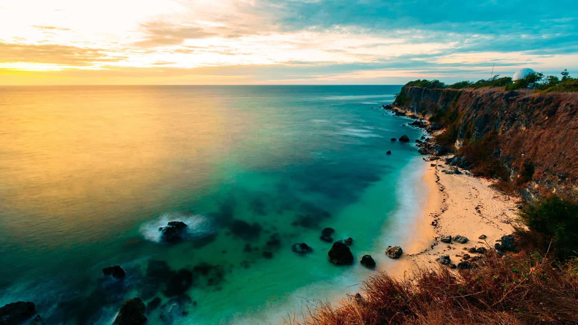 A stunning sunset over the ocean, illuminating the turquoise water beside a sandy beach and dramatic cliff near San Fernando.