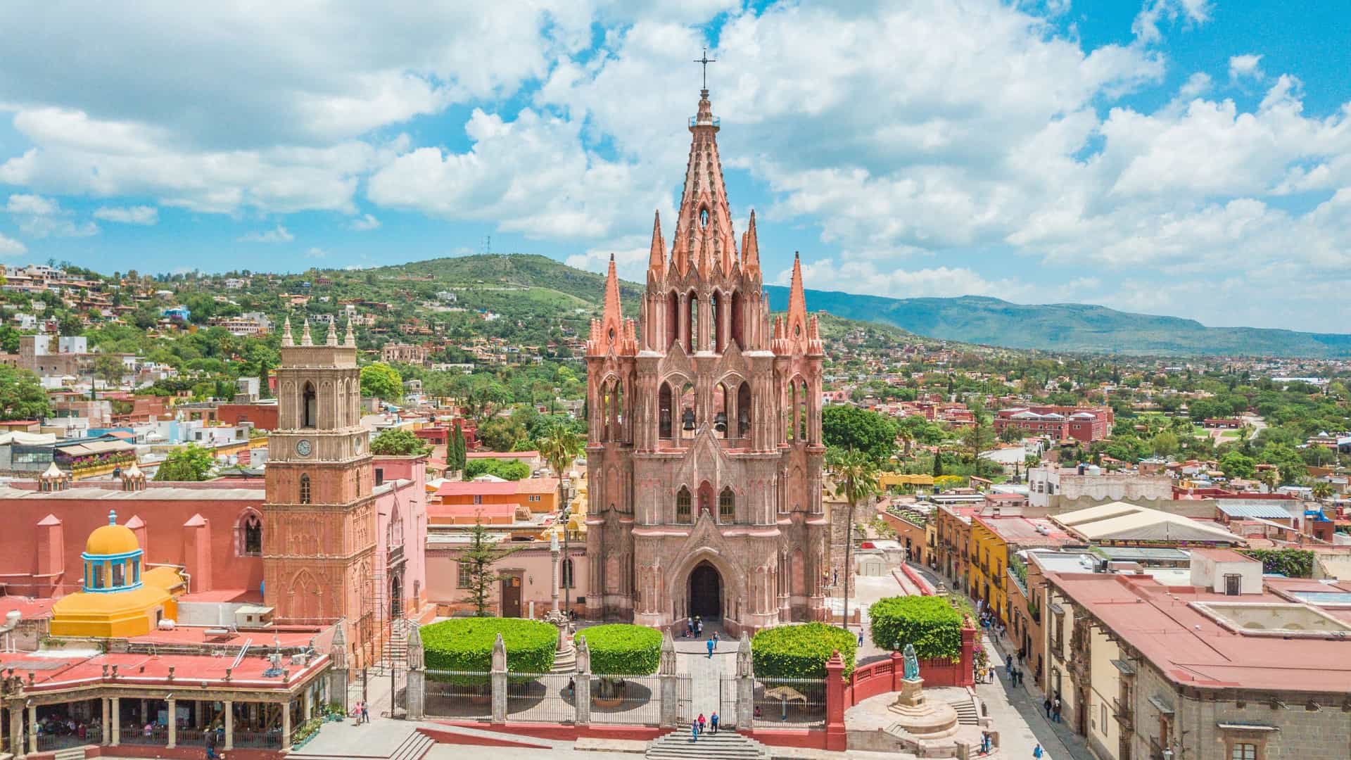 The pink stone neo-Gothic parish church, Parroquia de San Miguel Arcángel, dominates the city square under a cloudy blue sky.