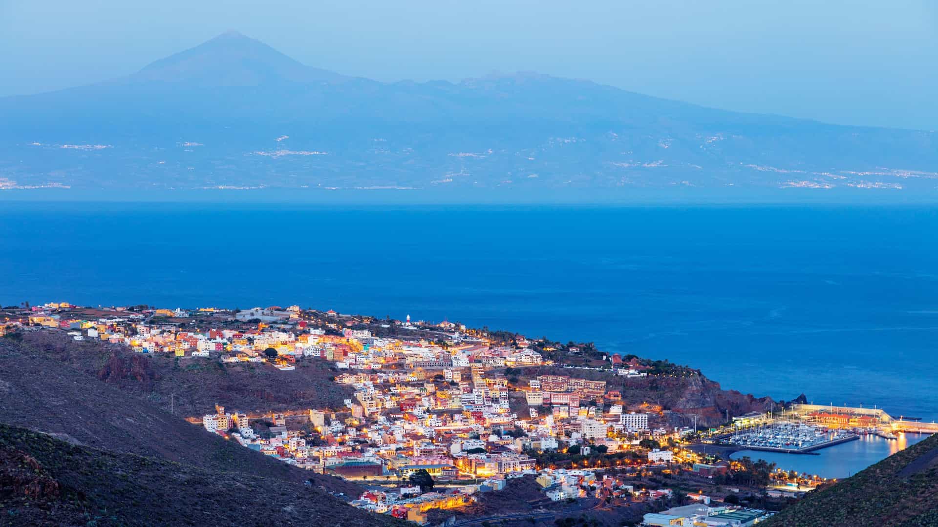 A twilight panoramic view of San Sebastian de la Gomera, Canary Islands, with the illuminated town and harbor in the foreground and Mount Teide of Tenerife in the distance.
