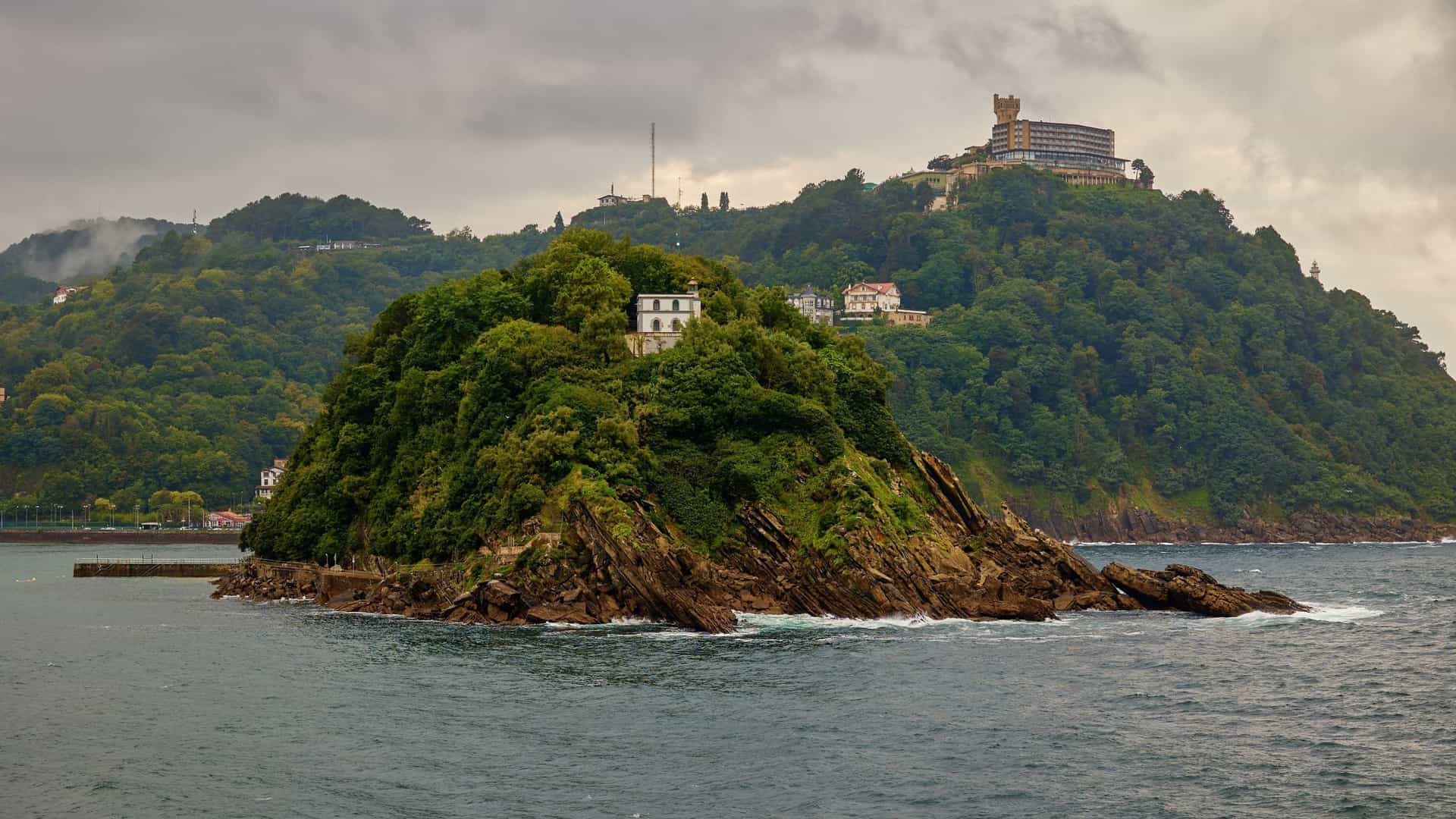 A lush, green island with a small white lighthouse sits in dark water, backed by a forested hillside with large buildings.