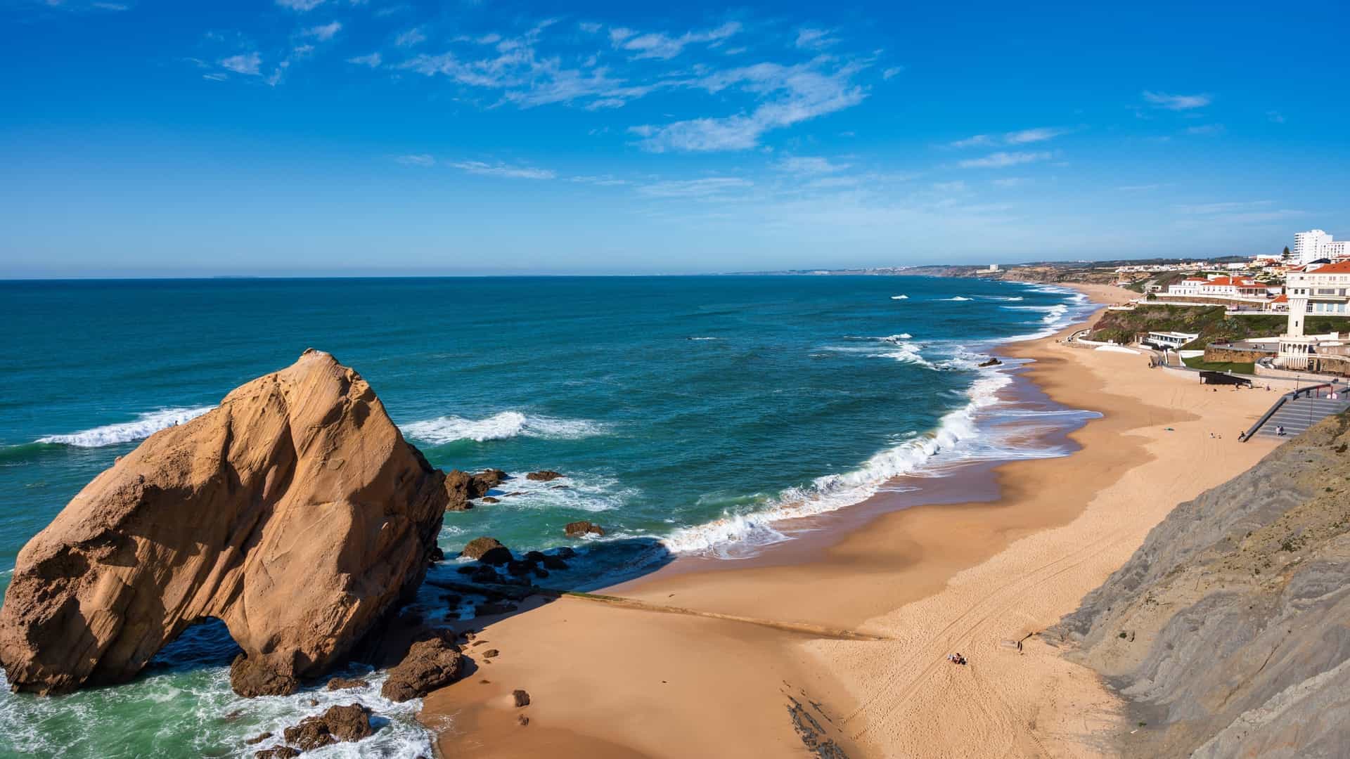 A scenic view of the Praia de Santa Cruz (Santa Cruz beach) in the municipality of Torres Vedras, Portugal, likely featuring a long stretch of sandy beach backed by cliffs or coastal rock formations, with the waves of the Atlantic Ocean rolling in. The image may prominently feature the distinctive Penedo do Guincho rock formation, a large boulder with a natural arch.