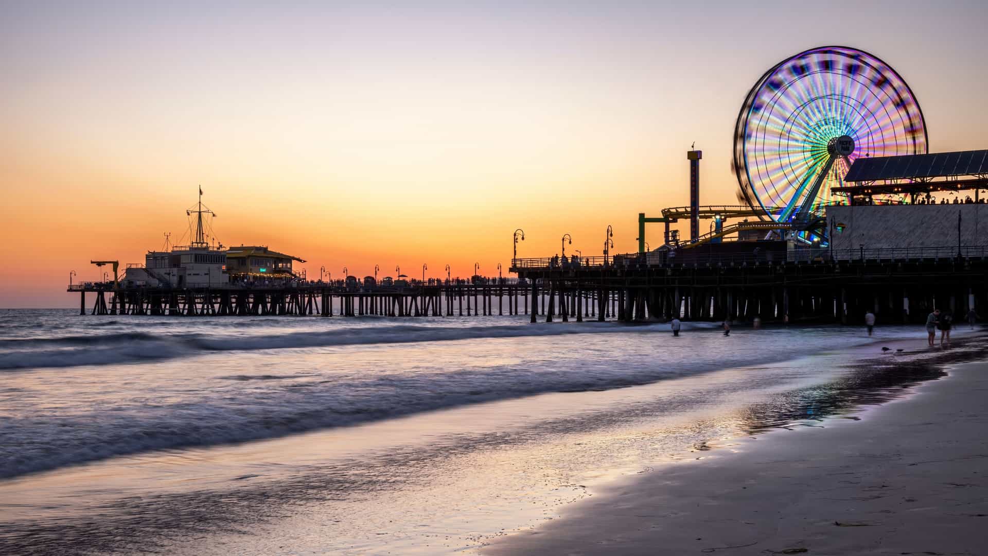The iconic Santa Monica Pier and its lit-up Ferris wheel are silhouetted against a vibrant orange and pink sunset over the ocean.
