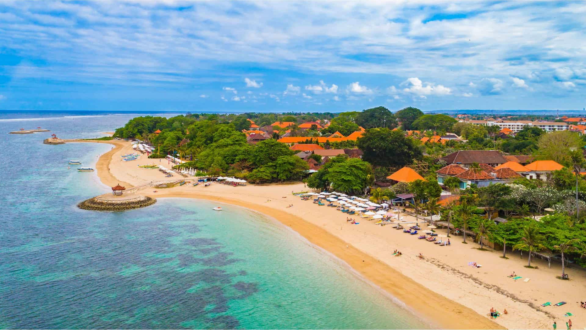 "An aerial view of the serene Sanur Beach in Bali, Indonesia, with a curved coastline, calm turquoise water, white sand, and beachfront resorts nestled in lush greenery."
