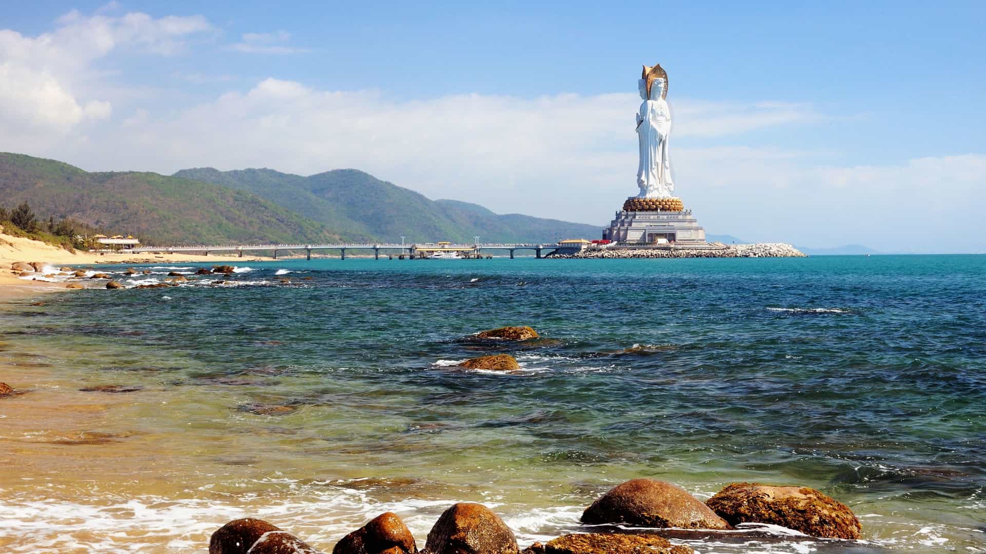 A large white statue of Guan Yin, the Goddess of Mercy, stands on a small island connected by a pier to the shore near Sanya, China.