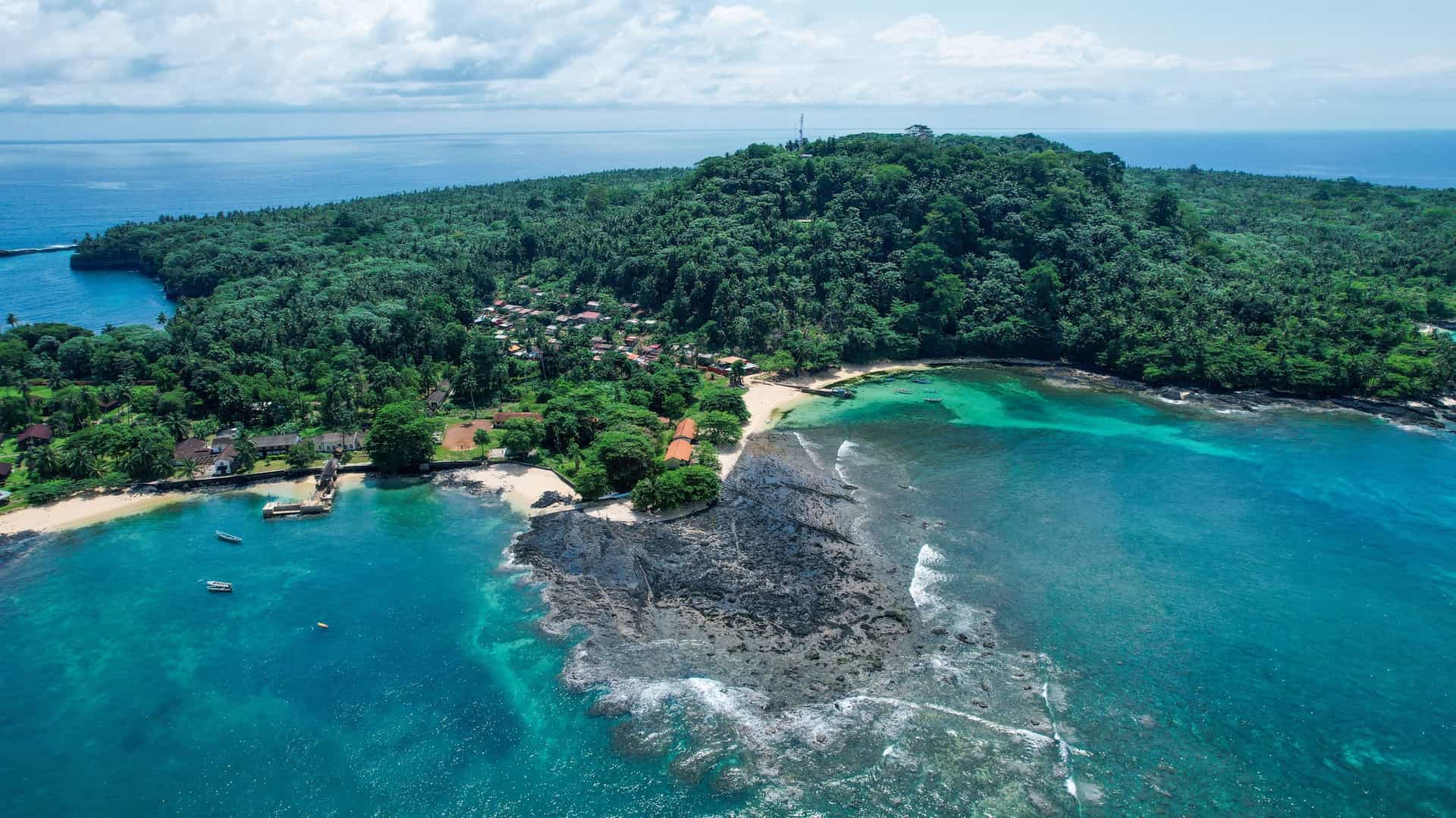 An aerial view of a remote beach and village on the tropical island of São Tomé, featuring lush green jungle and turquoise water.