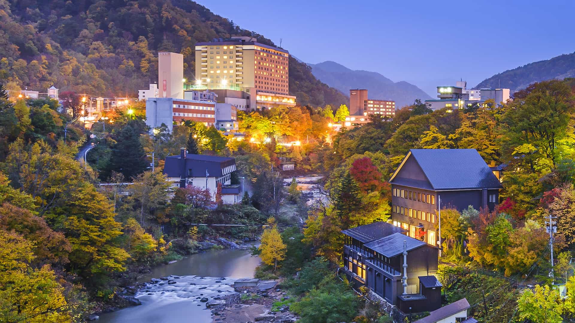 A serene dusk shot of Jozankei Onsen hot springs town in Sapporo, with a river flowing through a forested valley with buildings and hotels nestled on the hillsides.