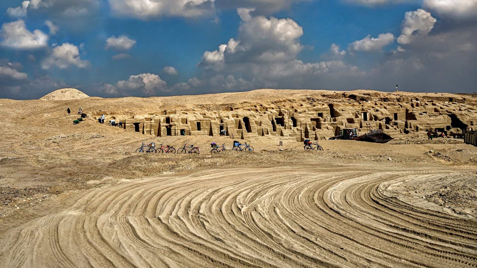 A curving dirt road leads toward several ancient, rectangular, rock-cut tombs built into a dry, tan desert hill in Egypt.