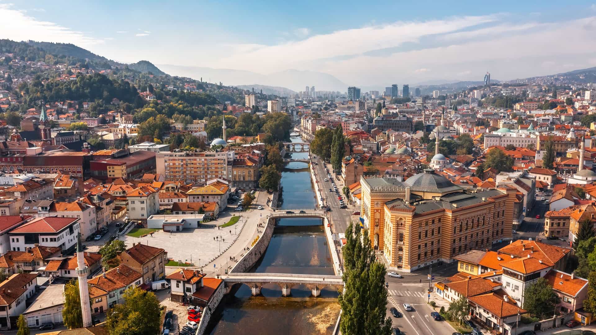 A high-angle view of the bustling city of Sarajevo, Bosnia and Herzegovina, with the Miljacka River flowing through the middle and surrounded by mountains.