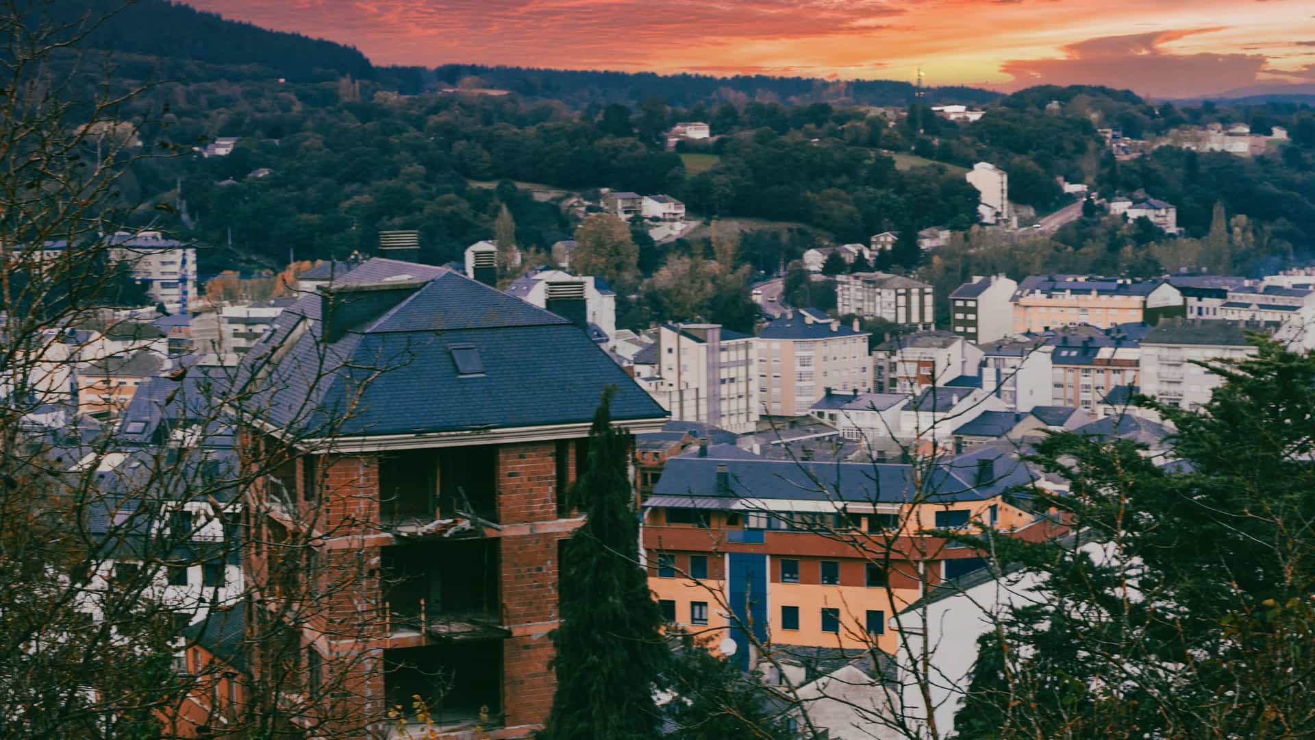 "A scenic, high-angle view of the town of Sarria, Spain, with its buildings and rooftops surrounded by a lush, forested landscape at sunset."