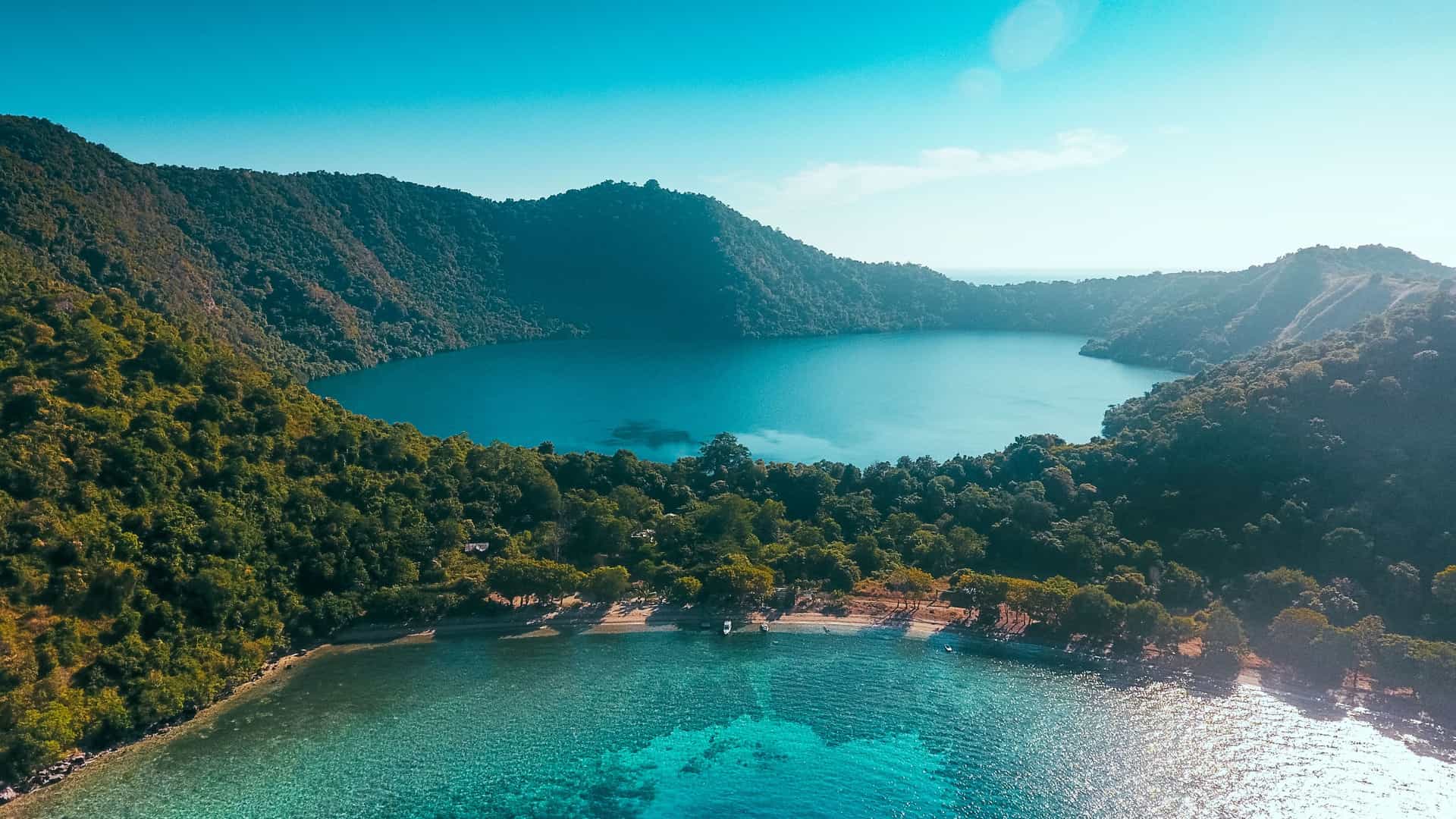 An aerial view of the volcanic Satonda Island in Indonesia, with a serene blue saltwater lake in its center surrounded by a lush green forest and the clear turquoise sea.
