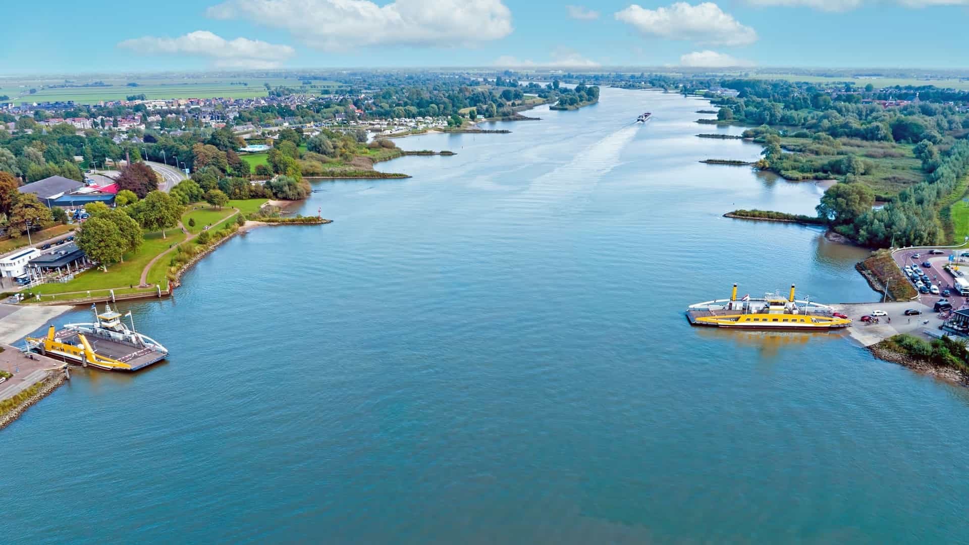 An aerial view of the wide, blue Lek River with two yellow car ferries crossing between grassy, developed banks.