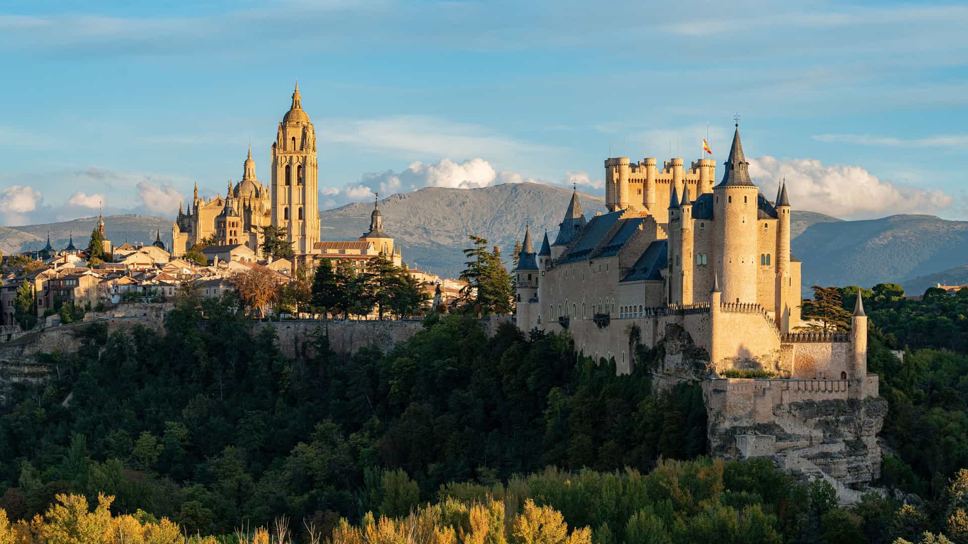 A wide-angle landscape view of the Alcázar of Segovia, a medieval castle, and the Segovia Cathedral towering over the city, with mountains in the background.