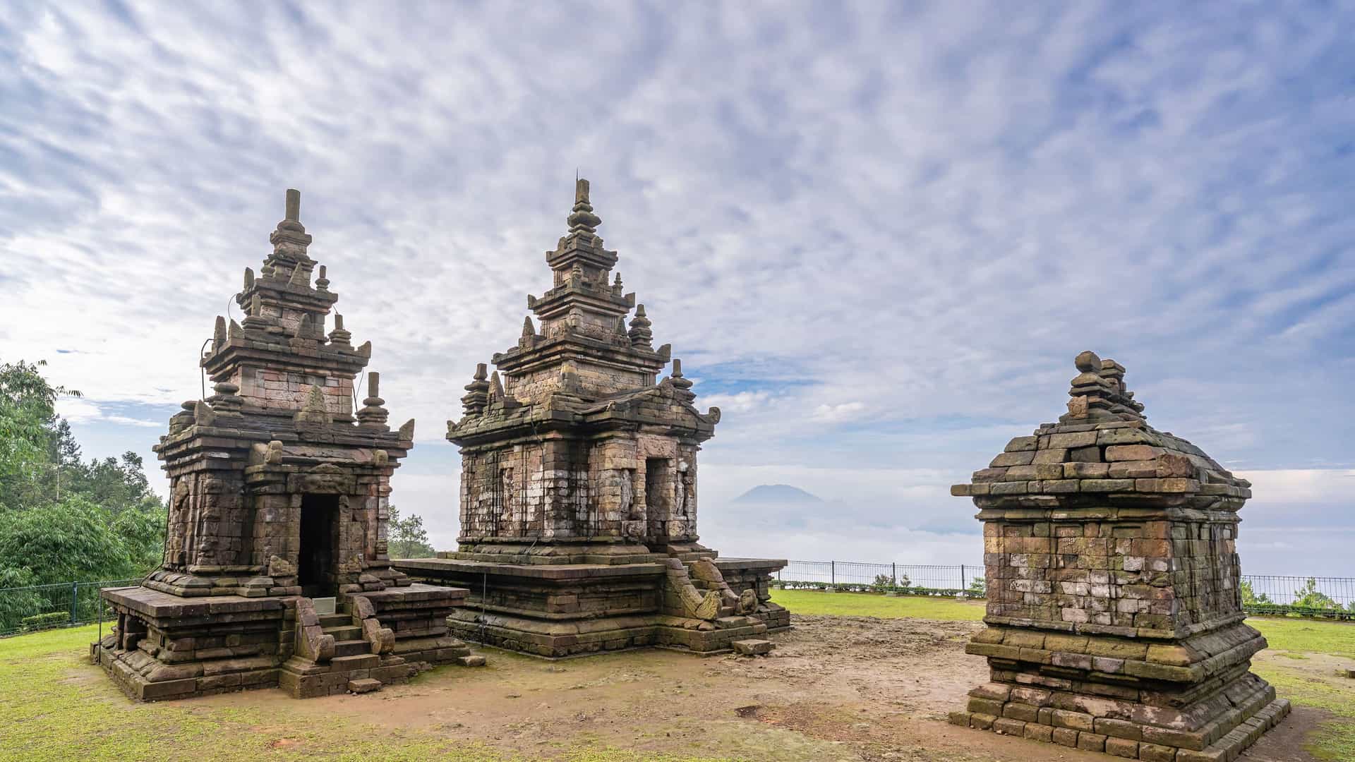 A stunning wide-angle view of the Gedong Songo temple complex, a collection of medieval Hindu temples on a misty hillside, with a mountain in the background in Semarang, Java, Indonesia.