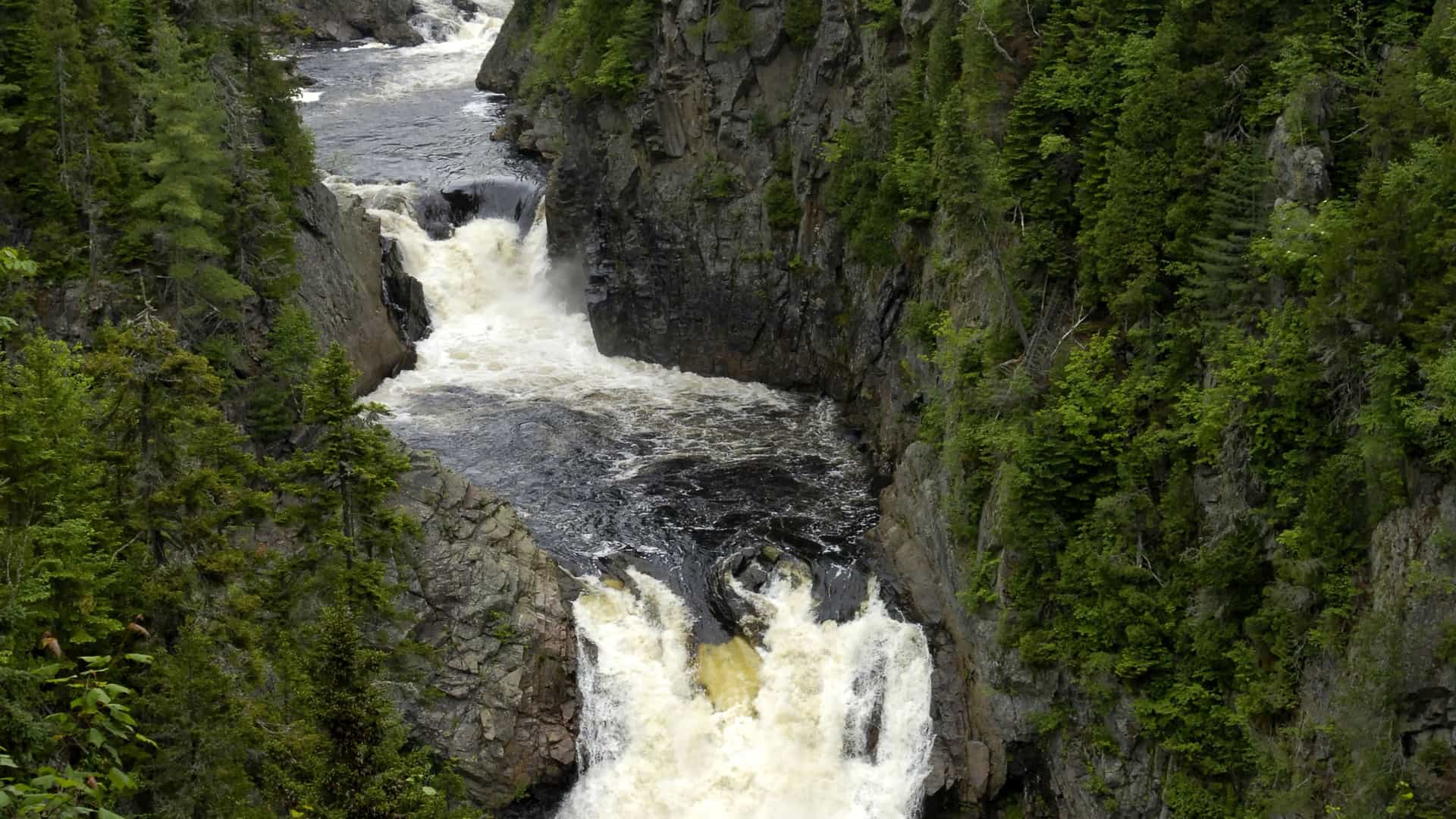 A dramatic view of the Sainte-Anne-du-Nord River cascading through a deep, rocky gorge surrounded by dense green forest near Sept-Îles, Quebec.