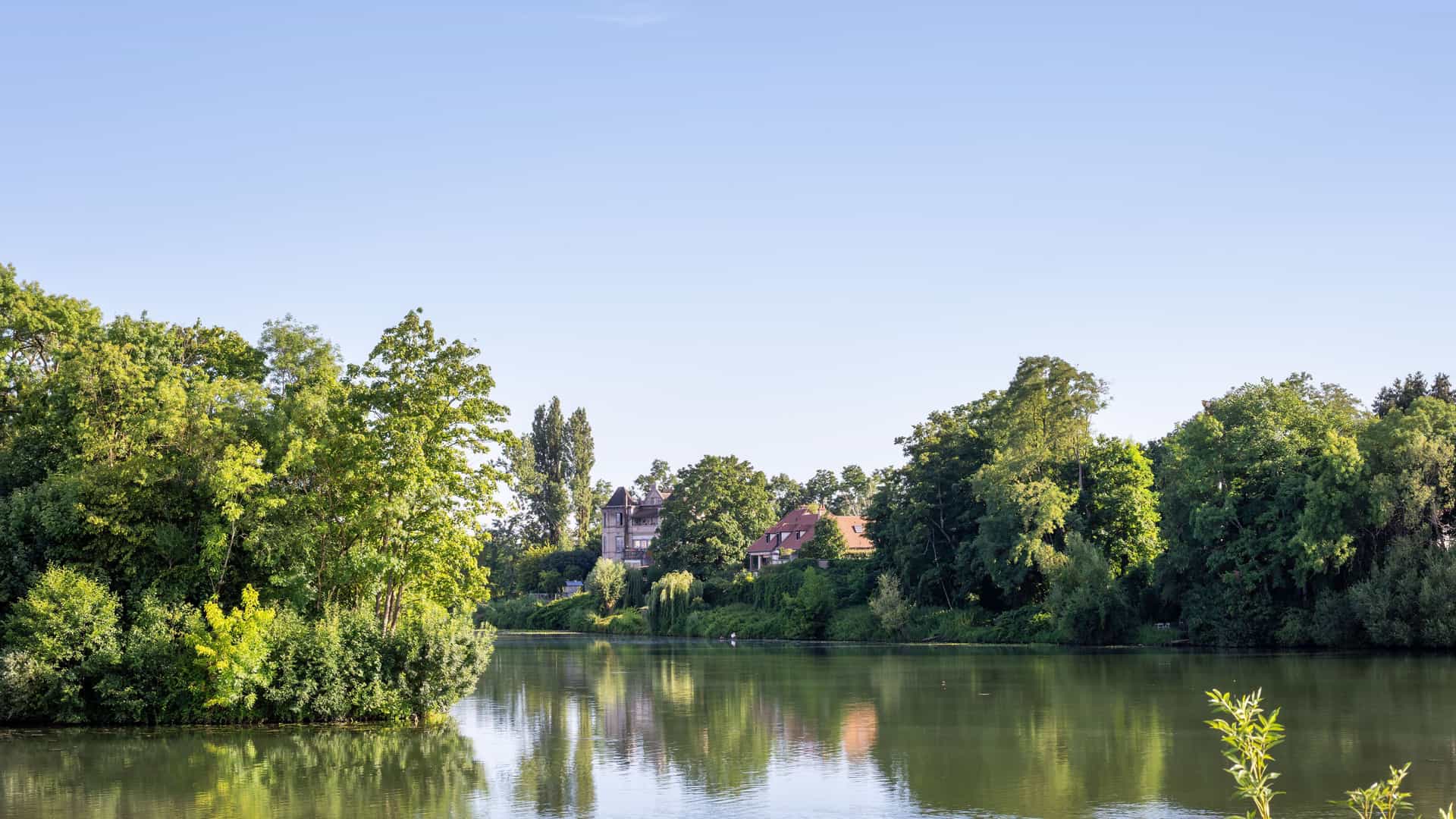 A tranquil wide-angle view of the Saône River in Seurre, France, with lush green trees lining the shore and historic houses reflected in the calm water under a clear blue sky.