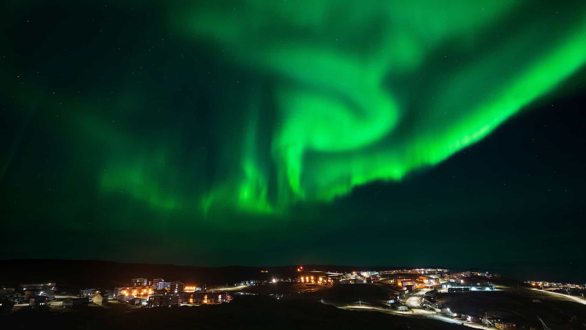 A wide-angle, low-light photograph of the aurora borealis, or Northern Lights, illuminating the night sky with vibrant green colors over a small village in Shaftesbury Inlet, Canada.