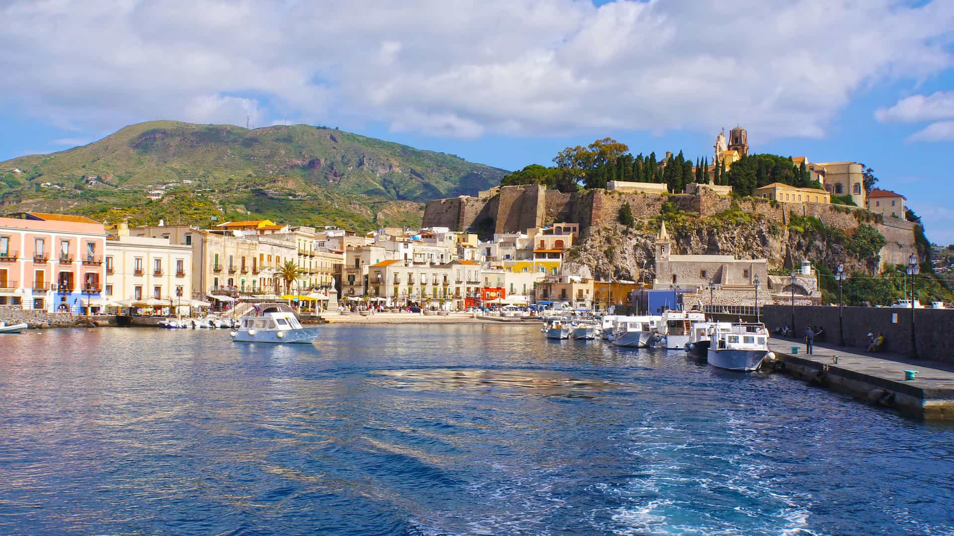 A picturesque view of Lipari, Sicily, with boats moored in the harbor and colorful buildings at the waterfront, with the impressive castle complex on a cliffside overlooking the town.