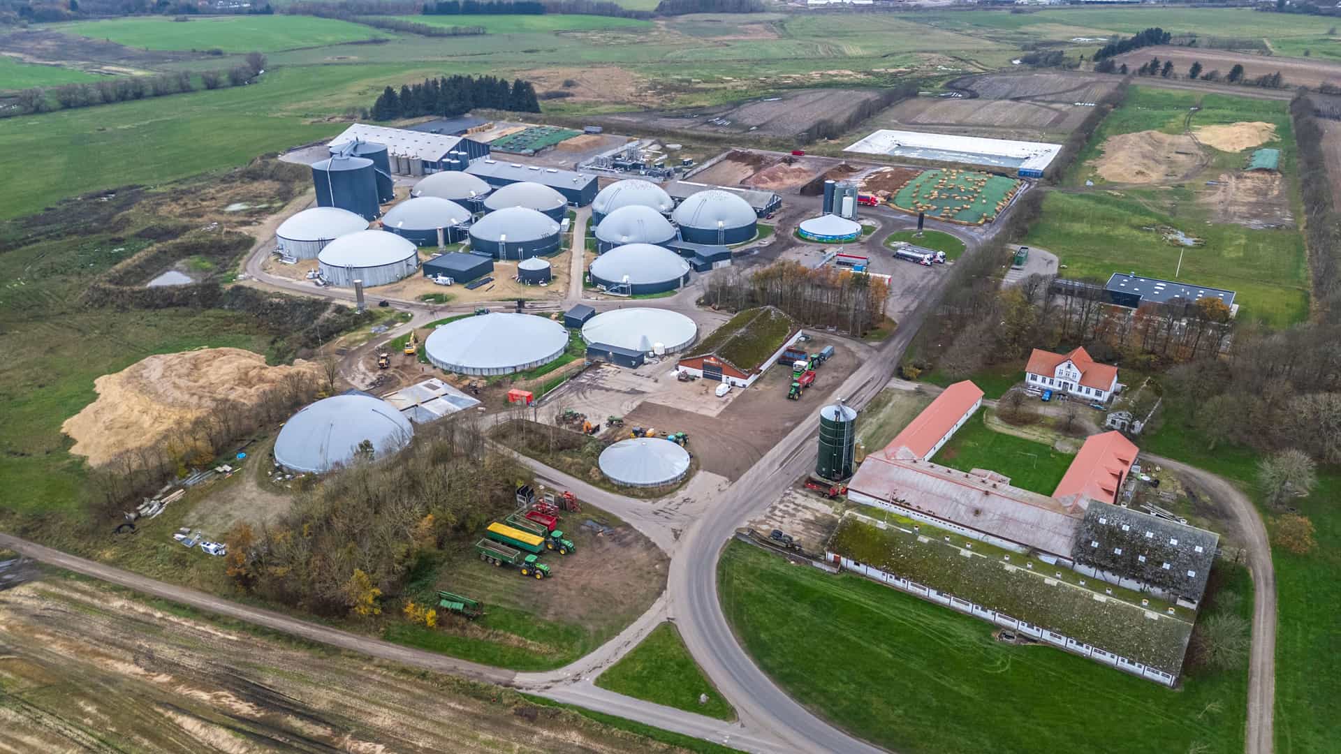 "An aerial view of a large, modern biogas plant with several domed storage tanks, surrounded by farmland and a traditional farm building in Sindal, Denmark.  "