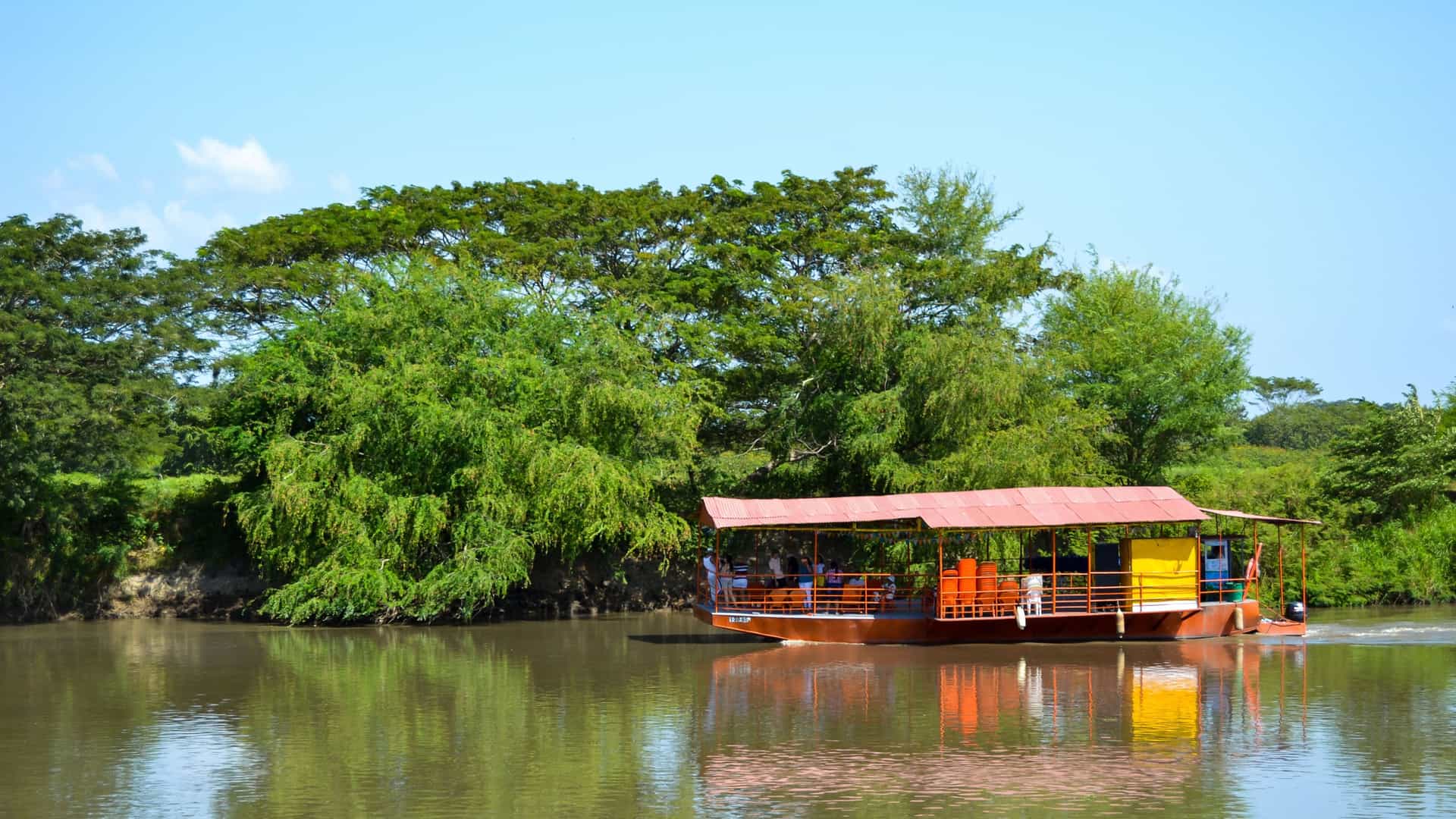 A riverboat with a red roof and seating for passengers travels along the calm Magdalena River, with lush green trees lining the banks of Sitio Nuevo, Colombia.