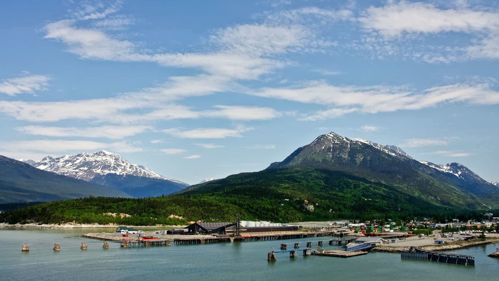 A beautiful view of the Skagway harbor and town with a long wooden pier and lush green forested mountains with snow-capped peaks in the background.