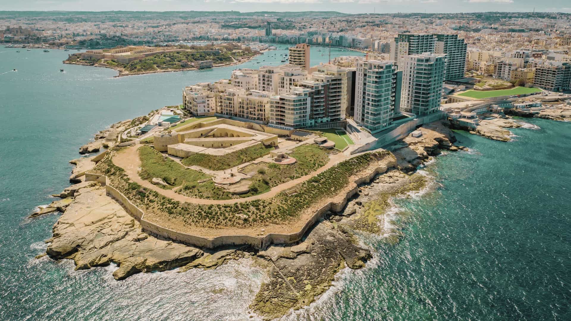An aerial view of the densely built coastal city of Sliema, Malta, focusing on a concentration of limestone buildings with traditional Maltese enclosed balconies and modern, multi-story developments. The clear, blue waters of the Mediterranean Sea or the Marsamxett Harbour would be visible in the background or along the waterfront.