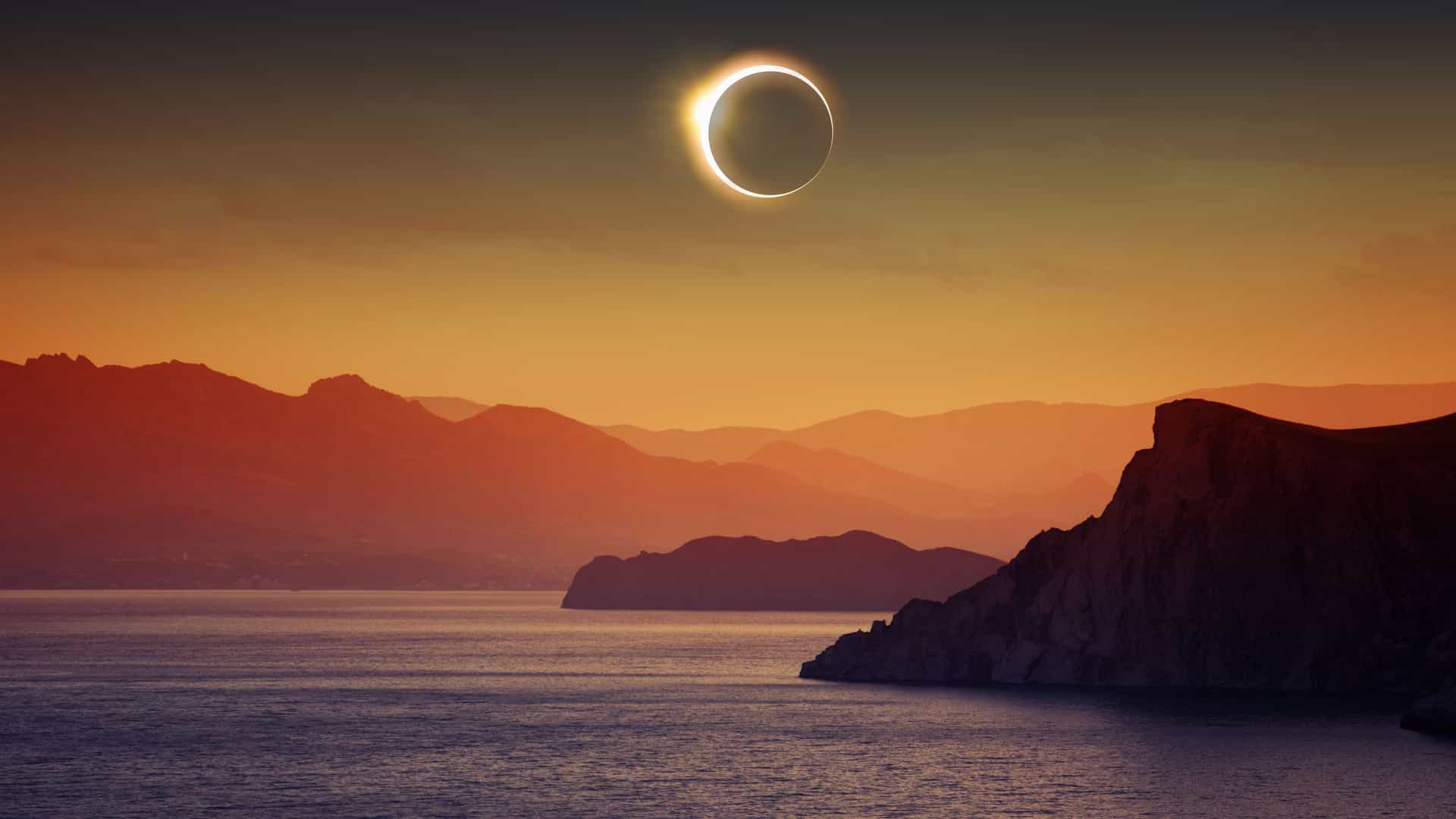 A dramatic celestial event over the sea with a ring of light, the total solar eclipse, visible over a tranquil body of water surrounded by silhouetted mountains.