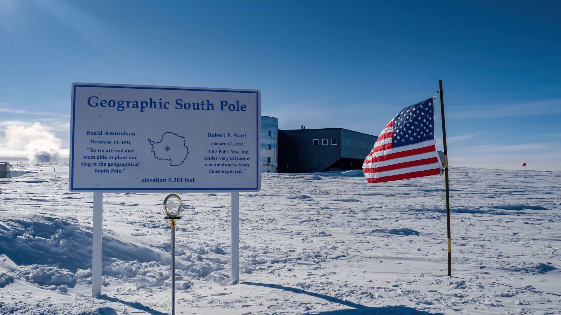 A scenic view of the Geographic South Pole marker and a large American flag waving in the wind, with the modern research building of Amundsen-Scott South Pole Station in the background.