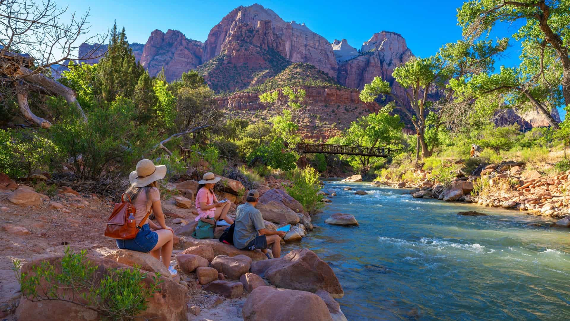 Tourists relax by the river, with a wooden bridge and towering reddish-pink cliffs of Zion National Park visible near Springdale, Utah.
