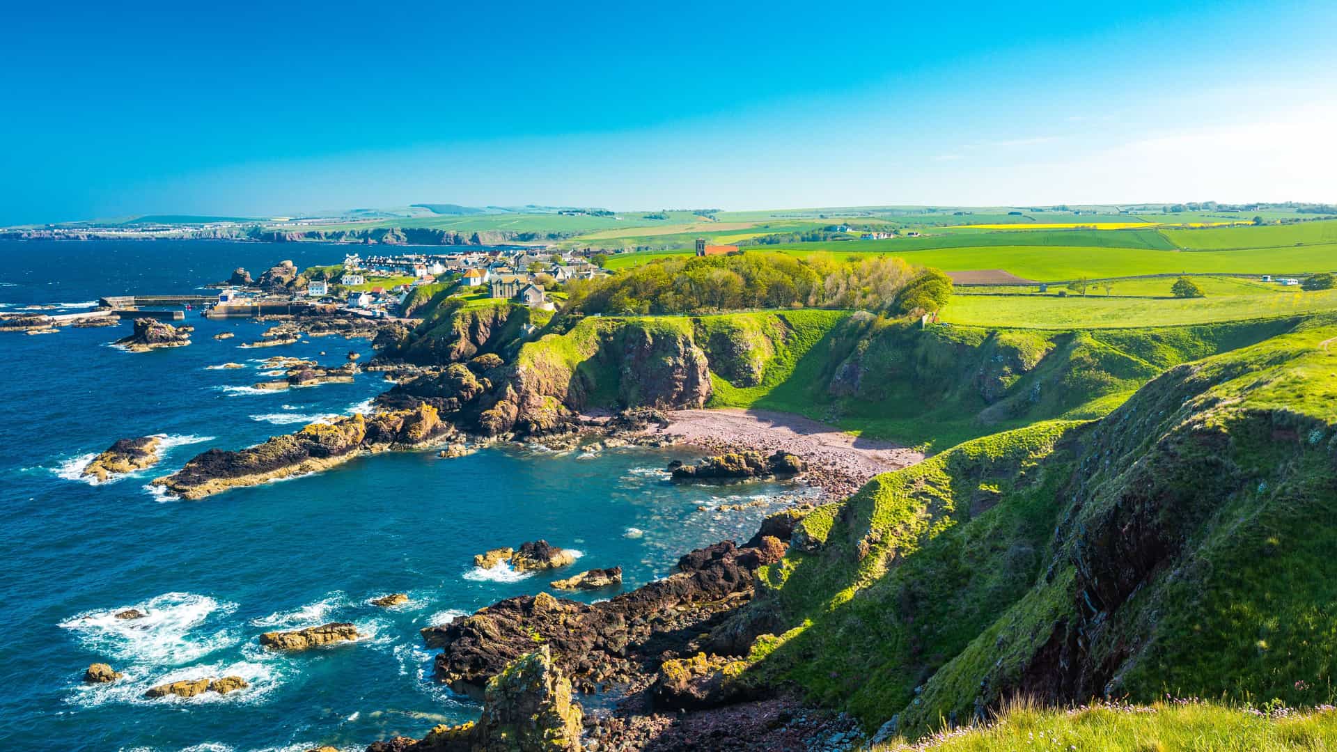 A panoramic view of the dramatic, rocky coastline of St. Abbs, Scotland, with the village nestled on a cliff overlooking the deep blue sea and lush green hills.