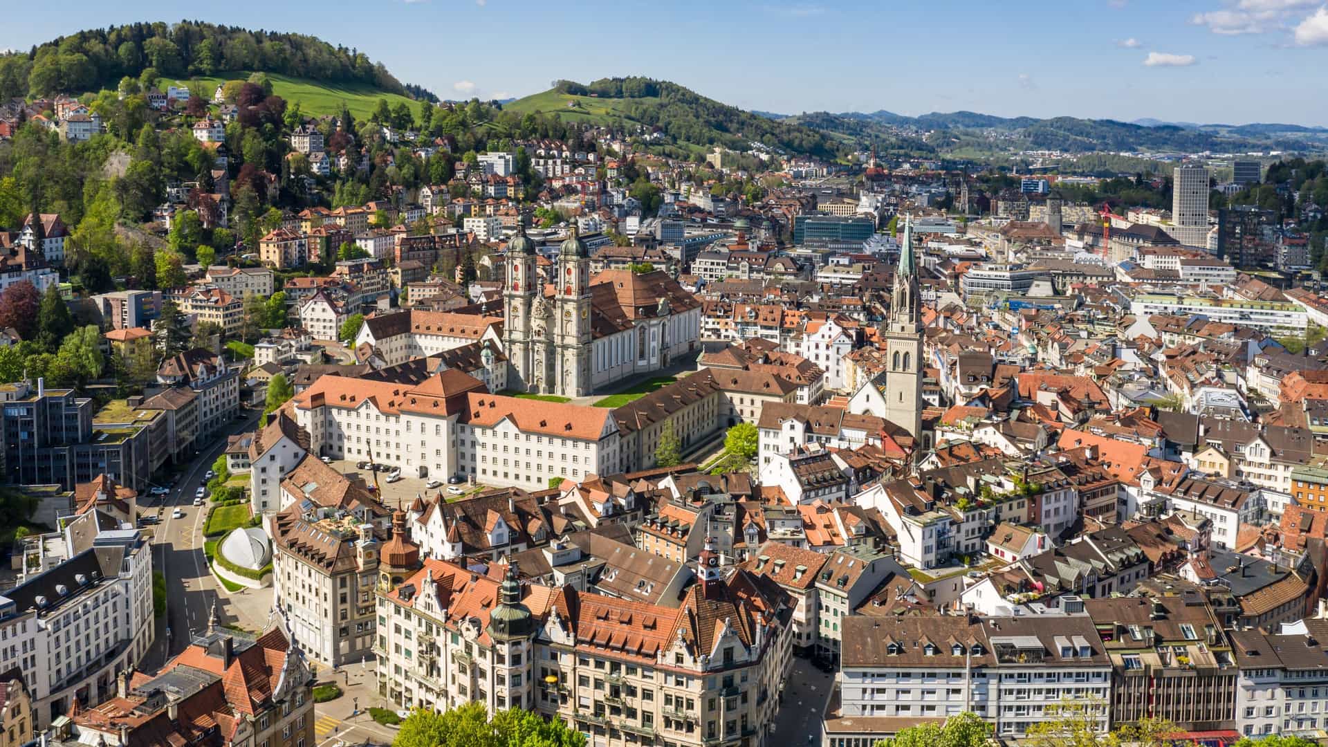 A high-angle aerial view of the historic Abbey of Saint Gall and its twin-towered cathedral, surrounded by the dense red-roofed buildings of St. Gallen, Switzerland, with rolling green hills in the distance.