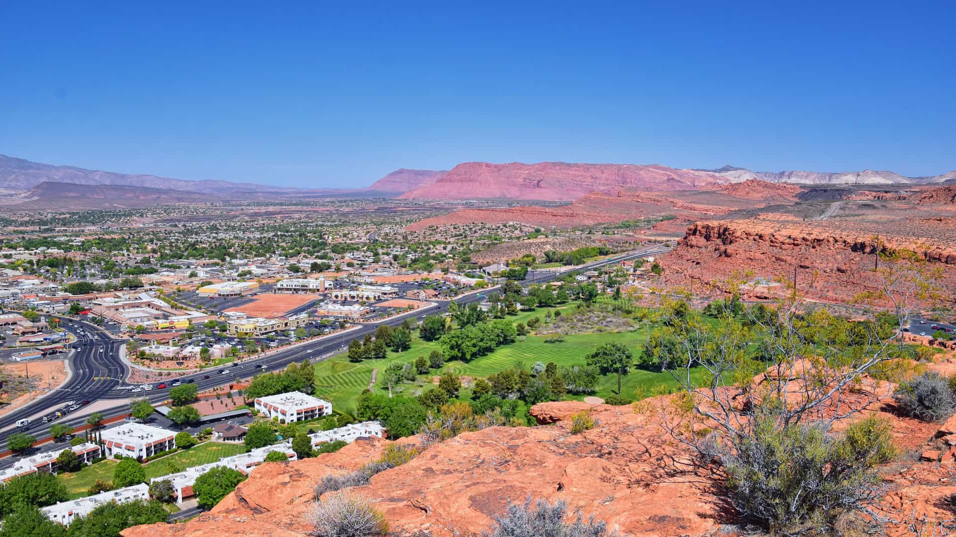 "A high-angle view of the city of St. George, Utah, with lush green golf courses and residential areas, surrounded by the dramatic red rock desert landscape and distant mountains under a clear sky.  "