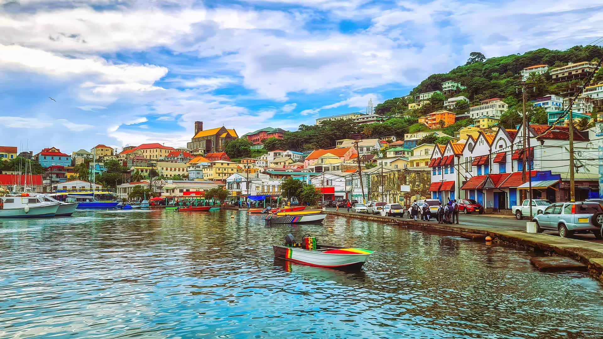A colorful hillside harbor in St. George’s Grenadines with vibrant buildings, a yellow‑roofed church steeple, calm reflective water, and boats lining the waterfront beneath a partly cloudy sky.