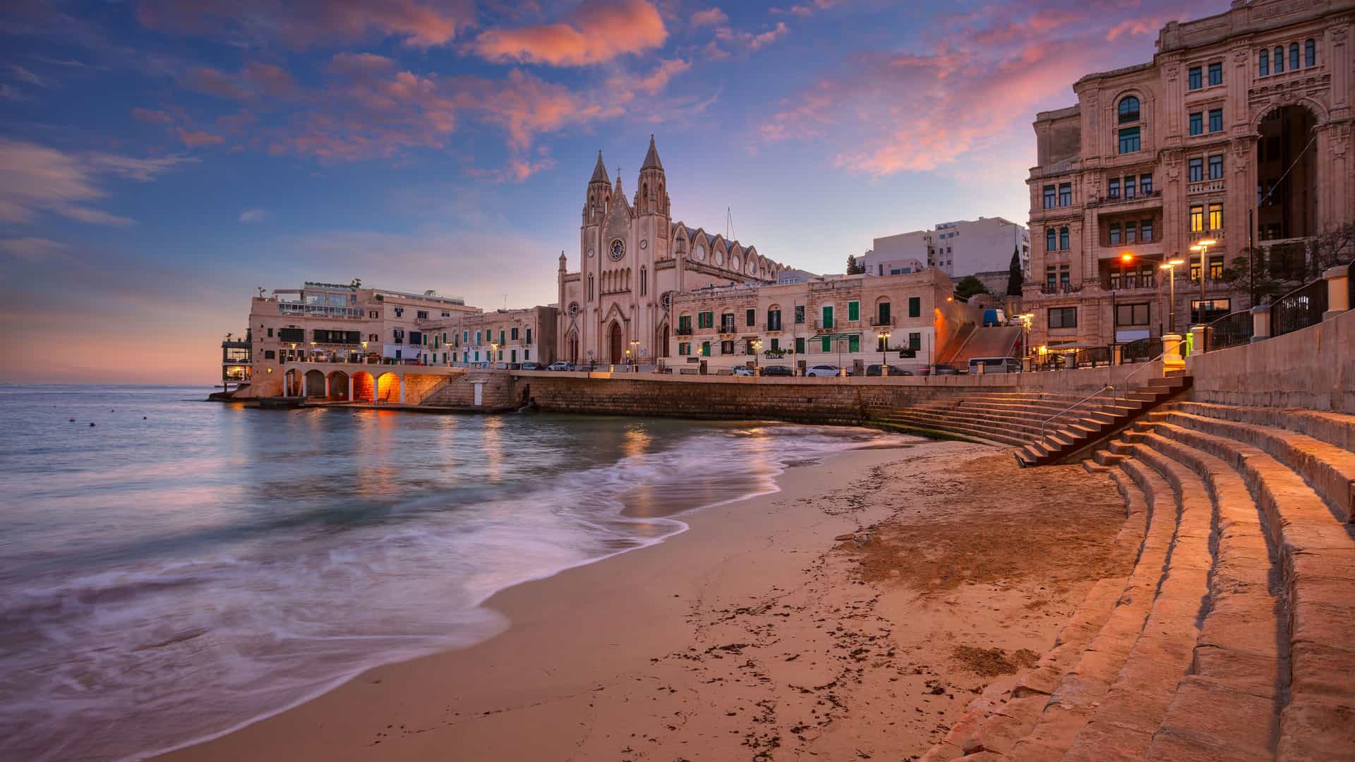 "A breathtaking sunset view of St. Julian's, Malta, with a sandy beach and gentle waves, showcasing the iconic Parish Church of Our Lady of Mount Carmel and the illuminated town buildings in the background.  "