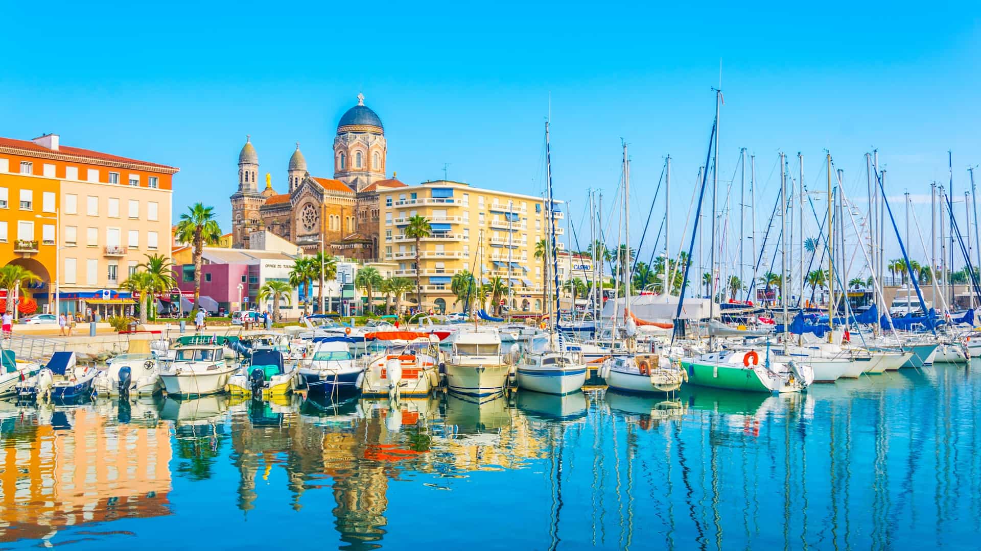 A scenic view of the marina in St. Raphael, France, with boats and yachts docked in the harbor, the Basilica of Notre-Dame de la Victoire, and colorful buildings in the background.