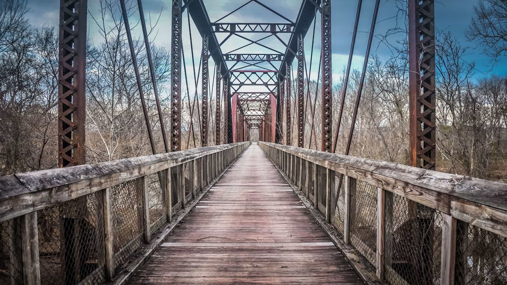 A low-angle view of a long, wooden walking bridge with a metal truss structure in Staunton, Virginia, surrounded by a forest of bare trees under a beautiful cloudy sky.