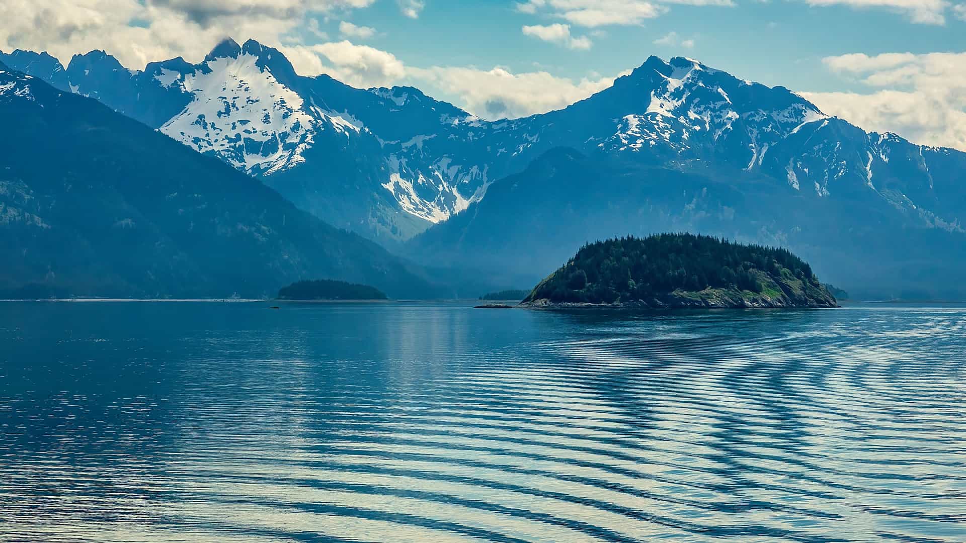 A beautiful panoramic view of the Stikine Strait in Alaska, with a tranquil, rippling blue sea, a small, lush green island in the middle, and a majestic range of snow-capped mountains in the background.