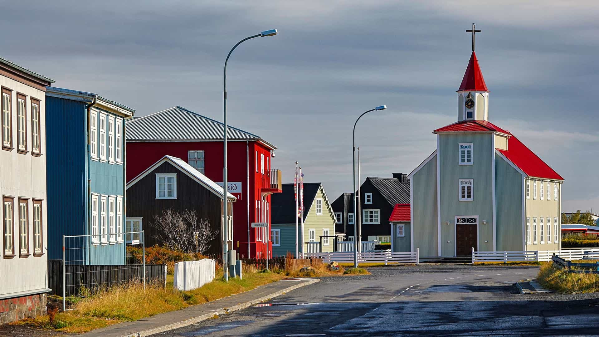 A colorful street scene in Stokkseyri, Iceland, with a red-roofed church and various multicolored wooden houses lined along a quiet street under a cloudy sky.