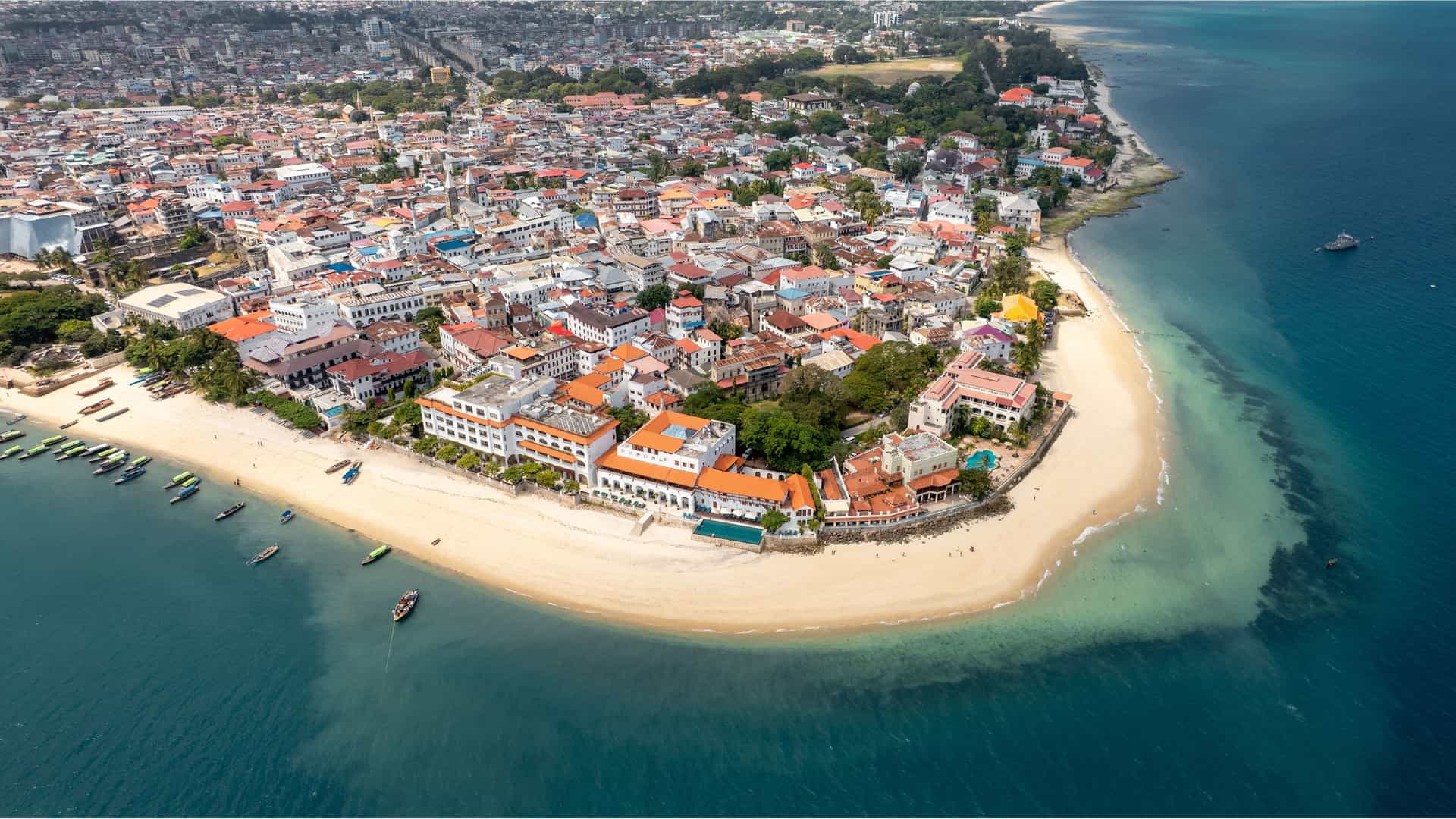 "An aerial view of Stone Town, a historic city in Tanzania, showing a dense cluster of buildings with red-tiled roofs along a pristine sandy beach next to the clear turquoise water of the Indian Ocean. "