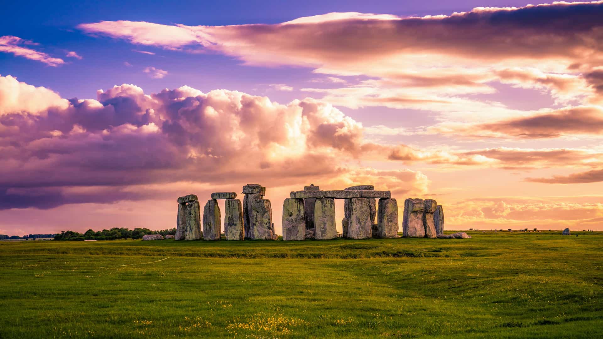 A wide-angle sunset view of the prehistoric Stonehenge monument in Wiltshire, England, with its iconic stone circle silhouetted against a dramatic purple and orange cloudy sky.
