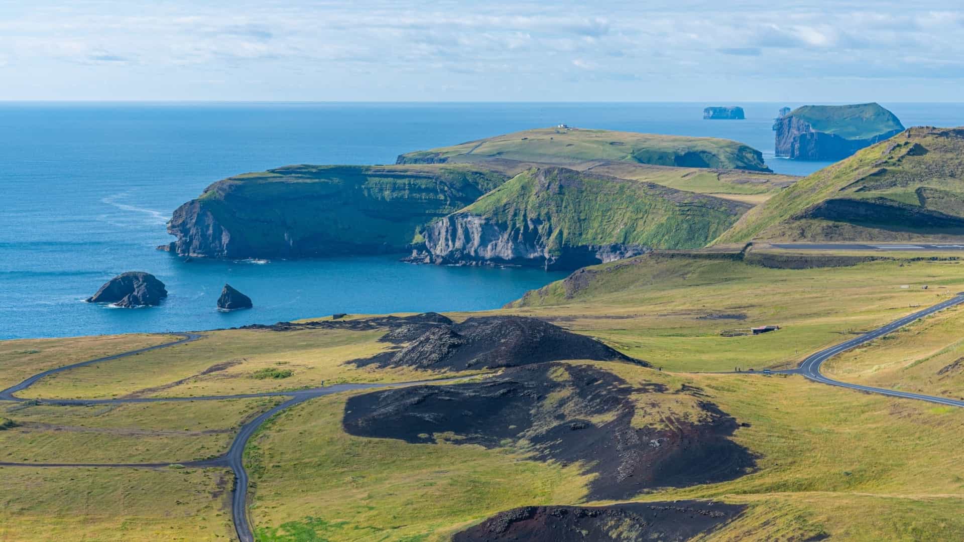 Grassy, rolling green and black hills meet the dramatic, rugged sea cliffs of Stórhöfði, with the blue ocean extending to the horizon.