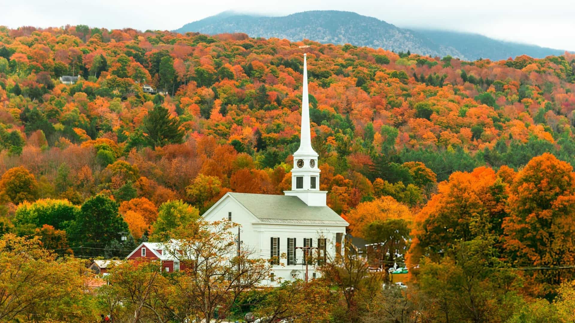 A white wooden church with a tall spire stands against a background of densely forested hills bursting with orange and red fall foliage.