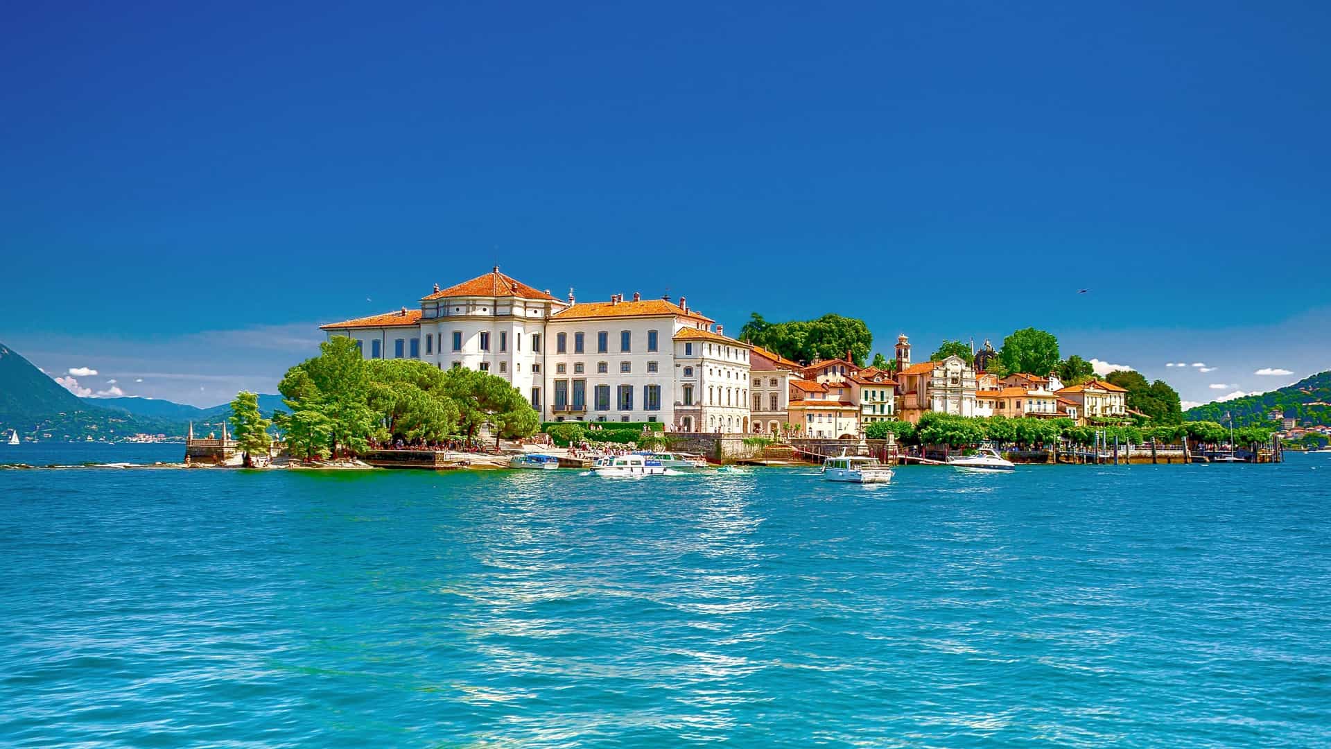A grand, white palace with a terra-cotta roof dominates a tree-covered island, Isola Bella, on the bright blue water of Lake Maggiore.