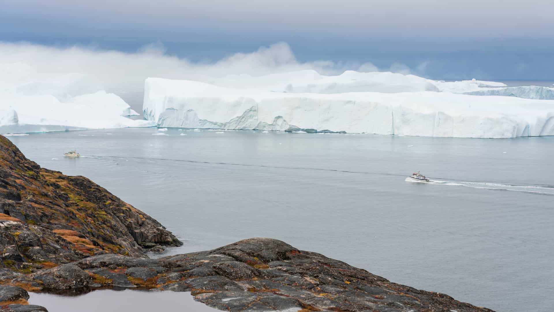 A dramatic Arctic scene in Stromfjord Nordre with towering icebergs floating in calm water, two boats navigating between them, and a rocky moss‑covered shoreline in the foreground beneath low mist and a pale sky.